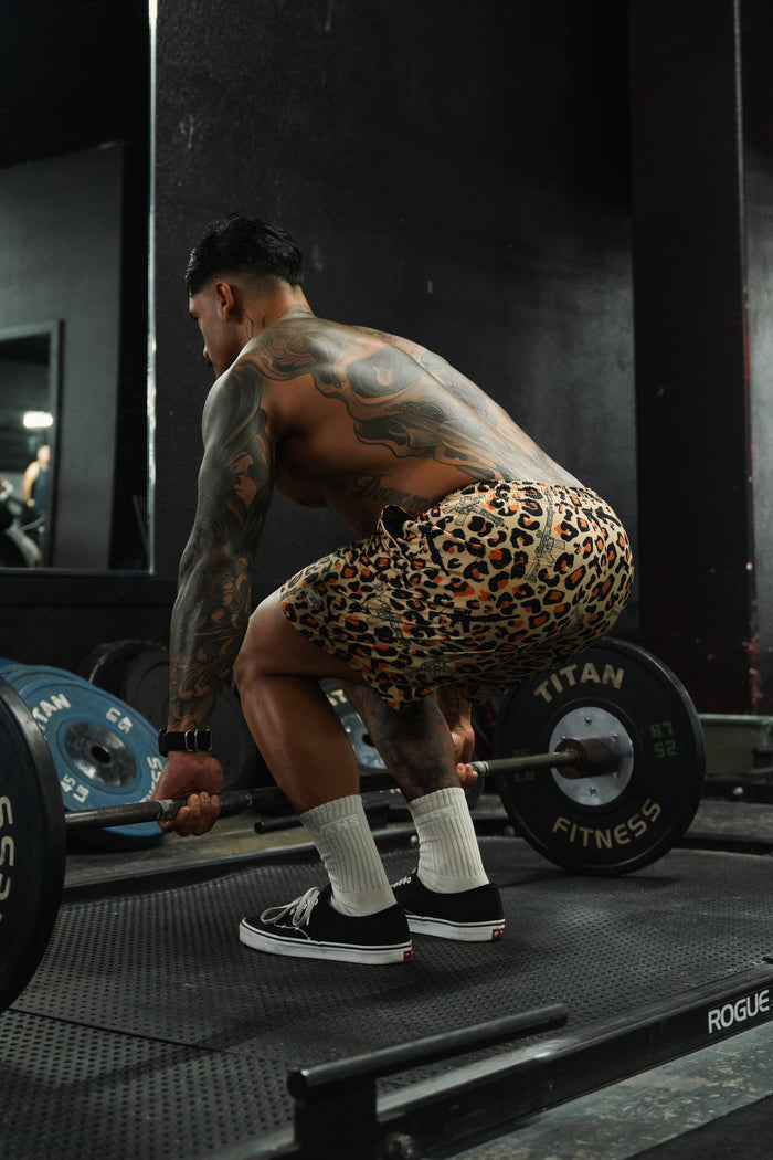 A muscular, tattooed individual wearing Tango Charlie Apparel Men's Performance Training Shorts | 5.5"- Loaded Leopard, white socks, and black sneakers squats in a gym, preparing to lift a barbell with weights.