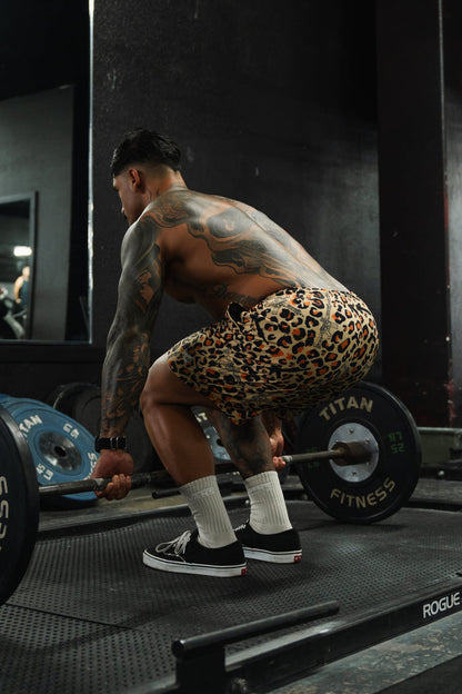 A muscular, tattooed individual wearing Tango Charlie Apparel Men's Performance Training Shorts | 5.5"- Loaded Leopard, white socks, and black sneakers squats in a gym, preparing to lift a barbell with weights.