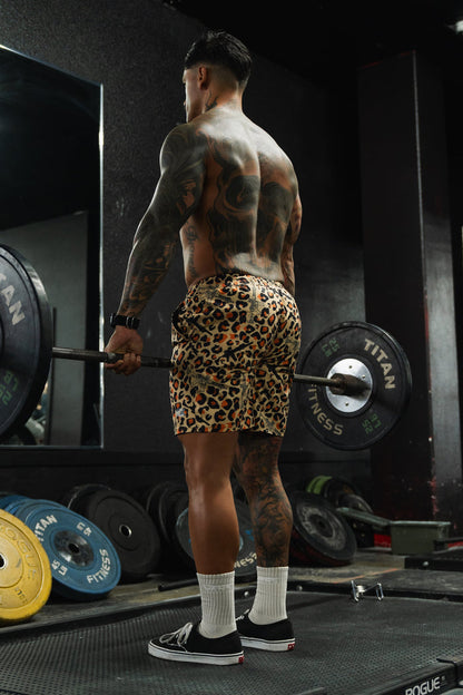 A muscular, tattooed man in Tango Charlie Apparel Men's Performance Training Shorts | 5.5"- Loaded Leopard and sneakers stands on a gym platform holding a barbell, with weight plates nearby and his reflection visible in the mirror.
