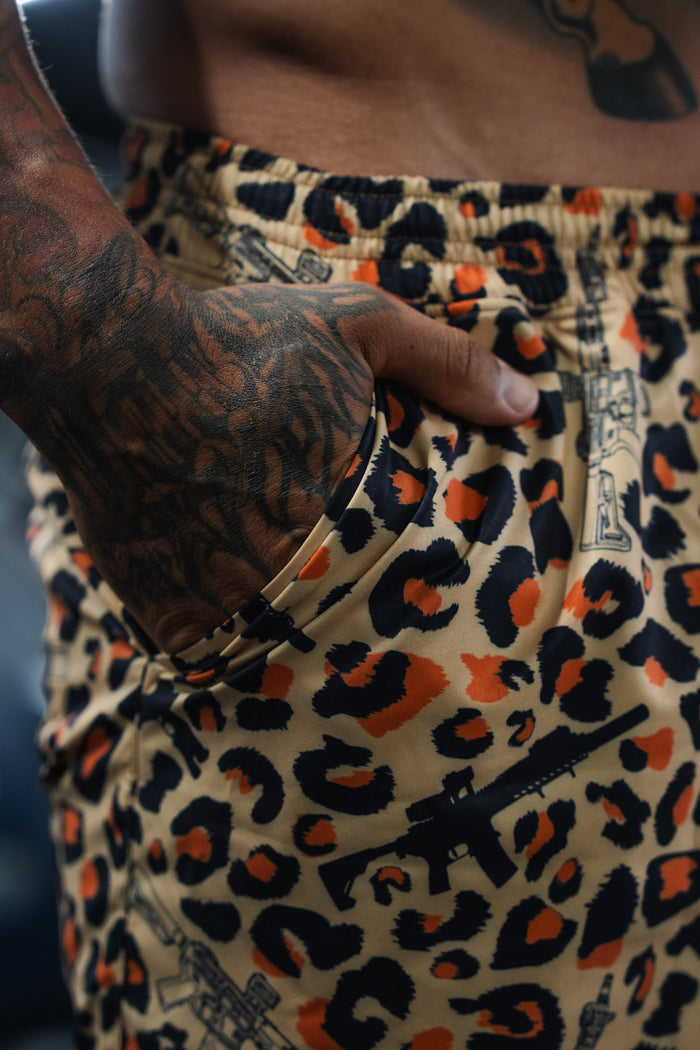 A close-up of a tattooed hand in the pocket of Tango Charlie Apparel’s Men's Performance Training Shorts | 5.5"- Loaded Leopard, which features a vibrant leopard print and black rifle graphics.