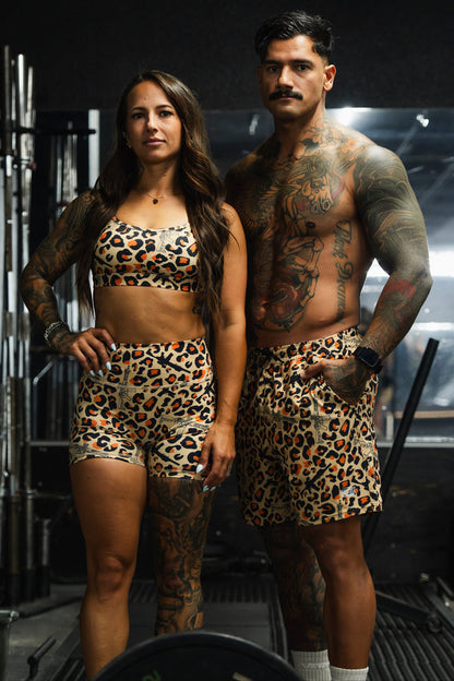 Two people at the gym show off tattoos side by side. The man wears Tango Charlie Apparel Men's Performance Training Shorts | 5.5"- Loaded Leopard, matching the woman's leopard print sports bra and shorts.