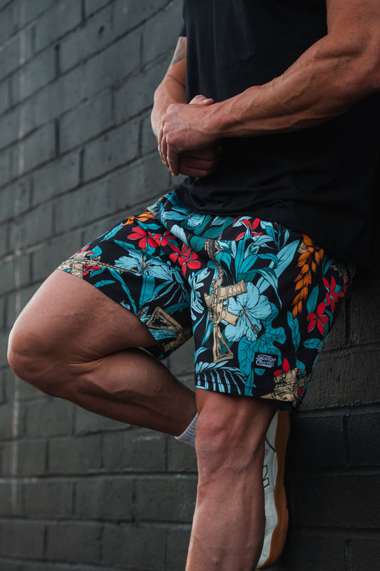 A person in a black shirt stands against a dark brick wall, wearing Tango Charlie Apparels Mens Performance Training Shorts in the Tactical Tropic pattern. The blue, red, and yellow leaves highlight their muscular arms as they elegantly pose with one leg bent.