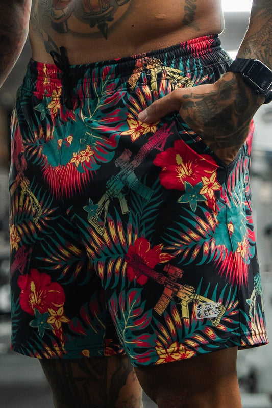 A tattooed man stands in a gym wearing Tango Charlie Apparel Men's Performance Training Shorts (5.5" - Tropical Assassin) and a smartwatch, with one hand in his pocket. Gym equipment is visible in the background.