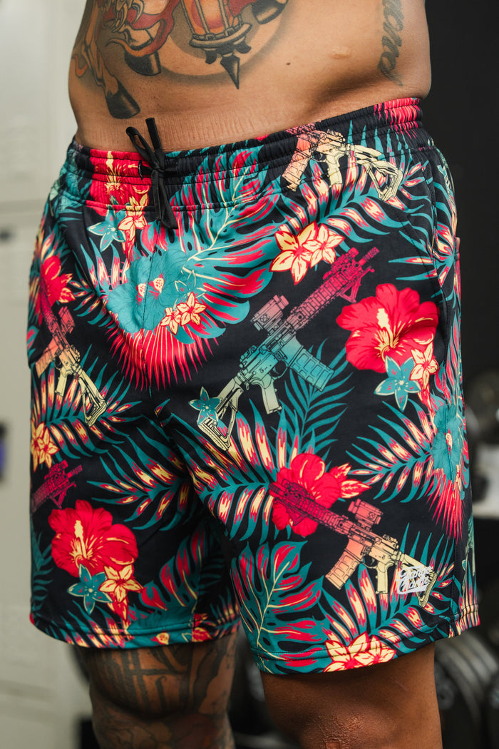 A person sports Tango Charlie Apparel's Men's Performance Training Shorts | 5.5" – Tropical Assassin, featuring vibrant floral and rifle prints. Visible tattoos decorate their torso and thigh—a true tropical assassin look.