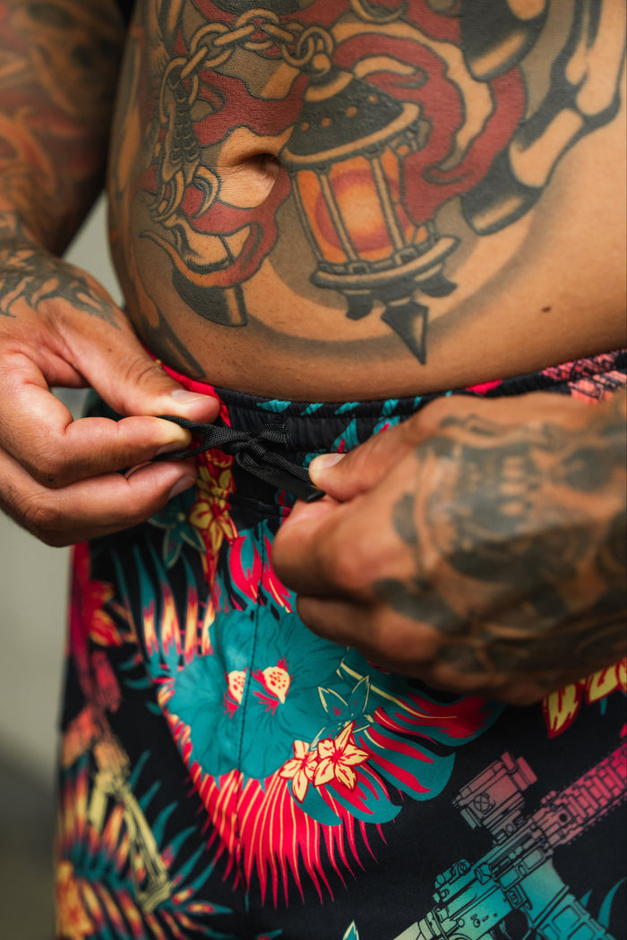 A close-up of tattooed hands adjusting the drawstring on Tango Charlie Apparel's Men's Performance Training Shorts | 5.5"—Tropical Assassin, with a lantern tattoo visible—perfect gear for standout tropical style.