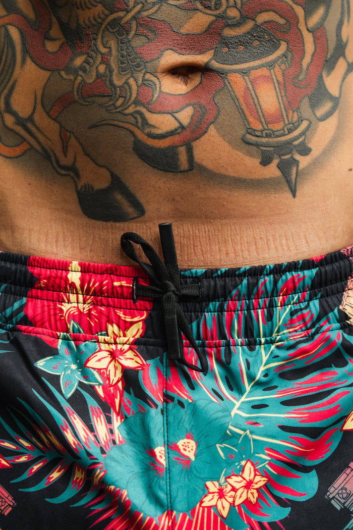 A close-up of a person's midsection shows colorful tattoos and vibrant Tango Charlie Apparel Men's Performance Training Shorts | 5.5"- Tropical Assassin featuring a floral pattern and black drawstring.