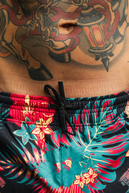 A close-up of a person's midsection shows colorful tattoos and vibrant Tango Charlie Apparel Men's Performance Training Shorts | 5.5"- Tropical Assassin featuring a floral pattern and black drawstring.