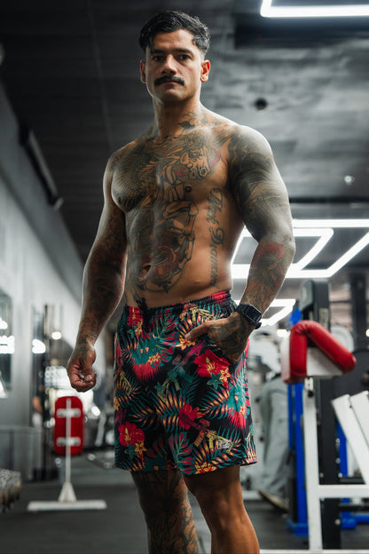 A muscular, heavily tattooed man with a mustache stands confidently in a neon-lit gym wearing Tango Charlie Apparel Men's Performance Training Shorts | 5.5" - Tropical Assassin, showcasing his bold training style among the equipment.