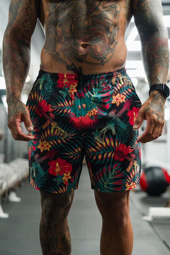 A muscular, tattooed man stands in a gym wearing Tango Charlie Apparel Men's Performance Training Shorts | 5.5" - Tropical Assassin, with workout equipment visible in the background.