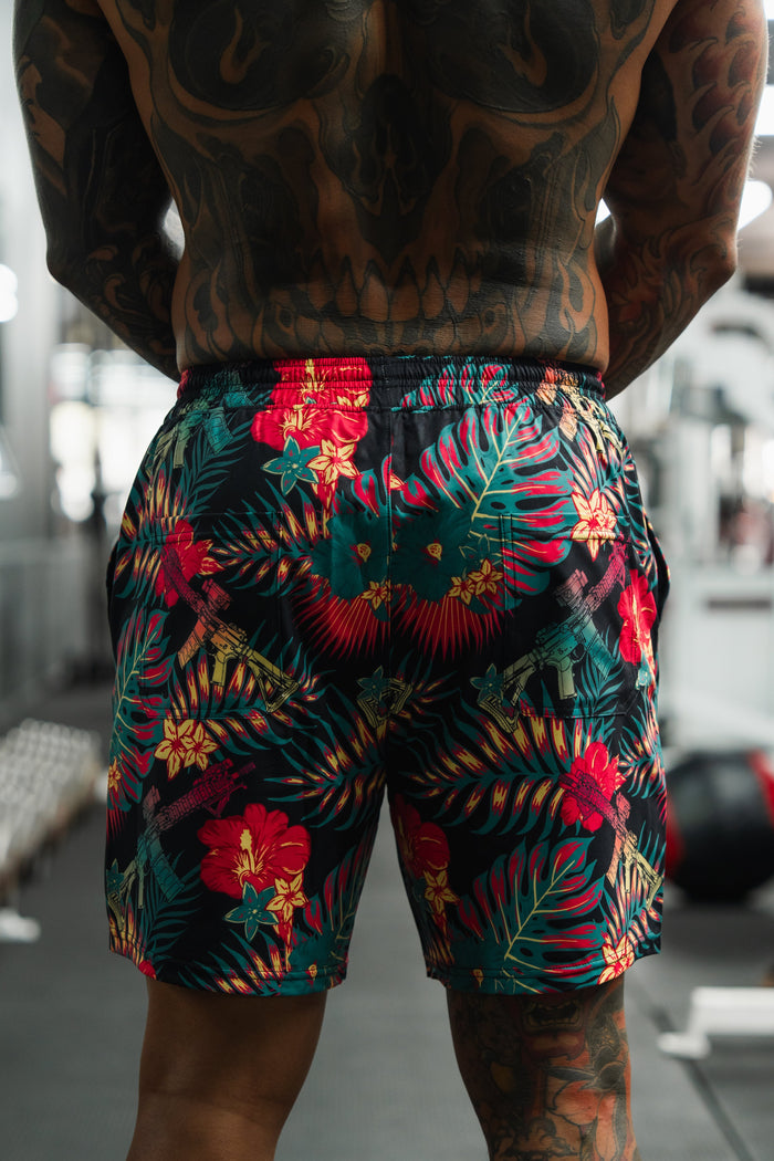 A tattooed individual stands in a gym wearing Tango Charlie Apparel's Men's Performance Training Shorts | 5.5" - Tropical Assassin, which feature a vibrant red and green floral design that draws attention to the lower back.