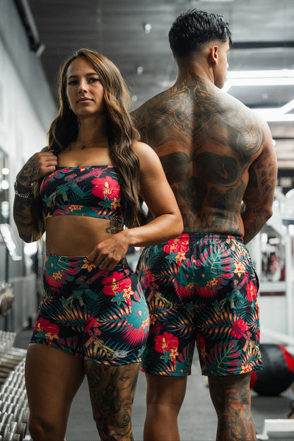 Two people with visible tattoos stand in a gym, wearing matching Tango Charlie Apparel outfits. The woman faces forward, while the man, back to the camera, sports Men's Performance Training Shorts | 5.5" - Tropical Assassin.