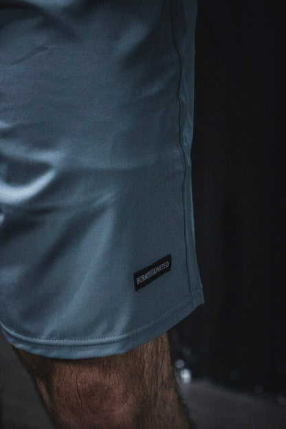 A close-up of a leg wearing Born United Private Label's Men's Standard Performance Shorts | 5.5"- Gunmetal Gray with a small tag near the hem against a dark, blurred background.