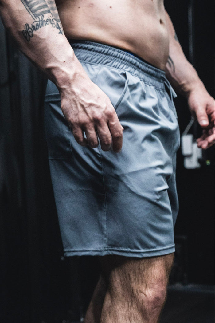 A close-up of a man's lower torso and upper legs shows him wearing Born United Private Label Men's Standard Performance Shorts | 5.5" in Gunmetal Gray, with his muscular, tattooed right arm by his side against a dark background.
