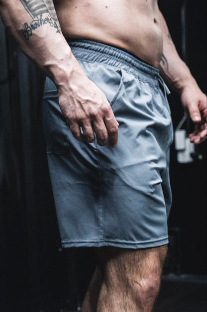 A close-up of a man's lower torso and upper legs shows him wearing Born United Private Label Men's Standard Performance Shorts | 5.5" in Gunmetal Gray, with his muscular, tattooed right arm by his side against a dark background.