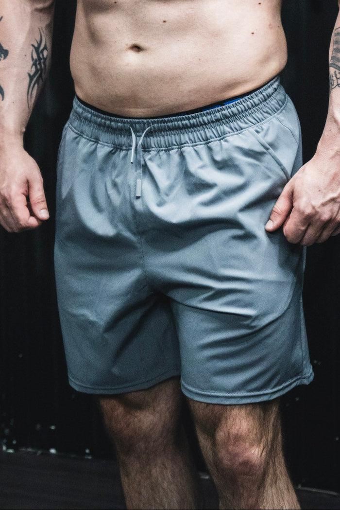 A person wearing Born United Private Label Men's Standard Performance Shorts | 5.5"- Gunmetal Gray stands with relaxed arms by their sides, upper body and head out of frame, and tattoos visible on their arms.
