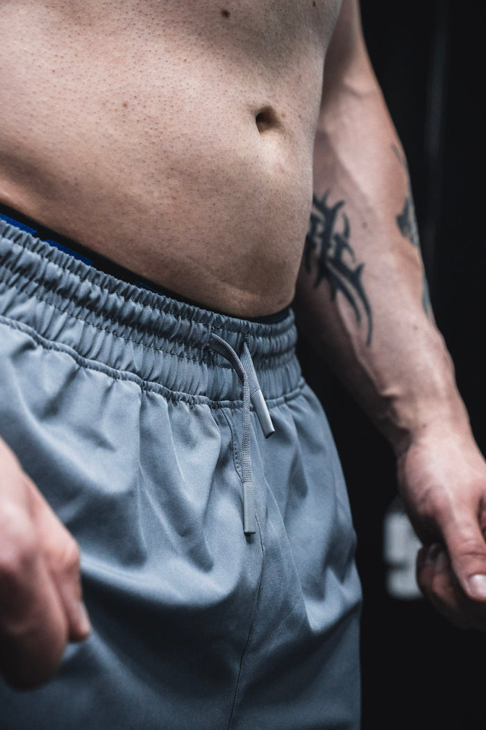 A close-up of a person’s bare torso and clenched fist, showcasing a tribal tattoo and wearing Born United Private Label Men's Standard Performance Shorts | 5.5" in Gunmetal Gray with a drawstring.