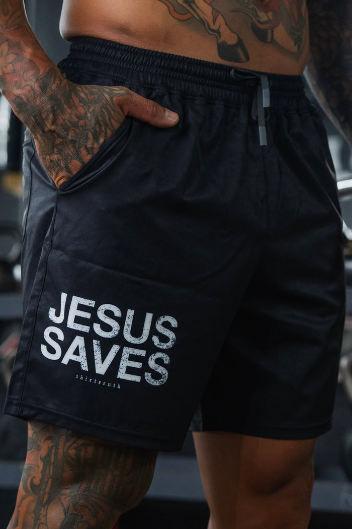 A tattooed individual wears 13th Men’s Standard Performance Shorts | 5.5"- Jesus Saves, featuring bold white lettering on the left leg. Their hand rests in their pocket, with a blurred background.