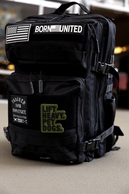 The Born United Private Label Midnight Black Premium Tac-Pac - 45L features tactical patches (American flag, BORN UNITED, TRAINED FOR VIOLENCE, LIFT HEAVY. PET DOGS) and is TSA-approved. Shown on a light-colored carpet indoors.