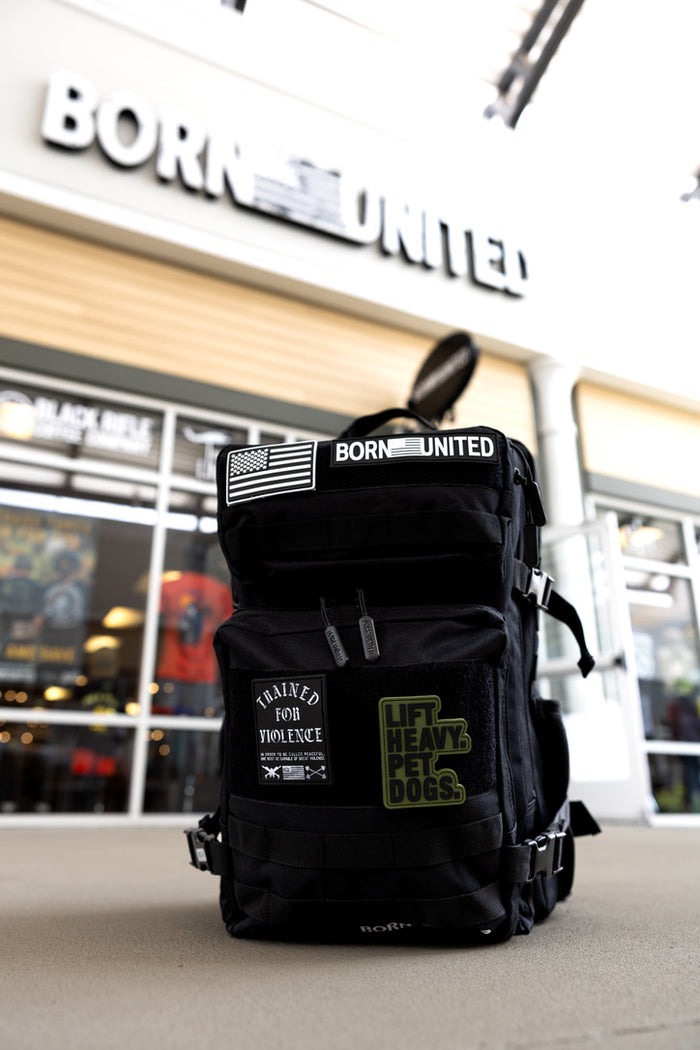 A Midnight Black Premium Tac-Pac - 45L from Born United Private Label, featuring "LIFT HEAVY. PET DOGS." and "TRAINED FOR VIOLENCE" patches, sits outside a store beneath a blurred Born United sign above the entrance.