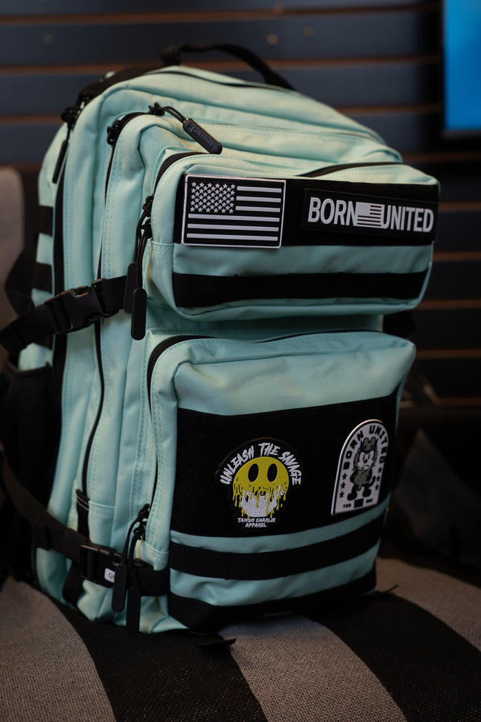 A Mint Condition Premium Tac-Pac - 45L by Born United Private Label, featuring a mint green and black design with multiple patches—including an American flag, "BORN UNITED," a skull graphic, and "UNLEASH THE SAVAGE"—rests on a striped chair.