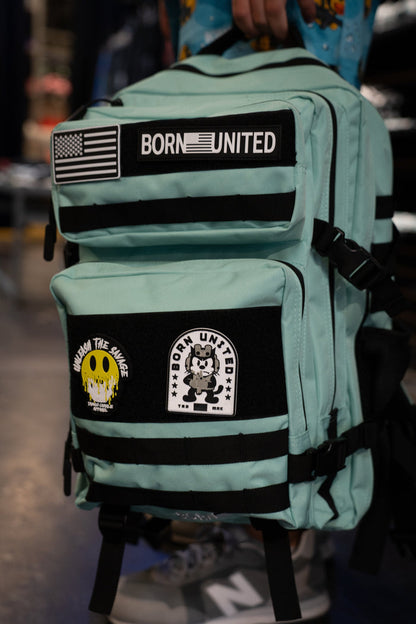 A person holds the Mint Condition Premium Tac-Pac - 45L by Born United Private Label, a mint green travel backpack adorned with three patches: an American flag with "BORN UNITED," a yellow smiley face, and a cartoon character patch also displaying "BORN UNITED.