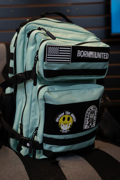 A Mint Condition Premium Tac-Pac - 45L travel backpack from Born United Private Label in light blue, featuring black zippers and decorated with patches such as an American flag, BORN UNITED logo, a yellow skull, and a cartoon animal, resting on a chair.