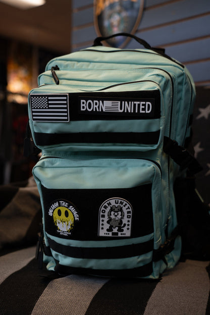 The Mint Condition Premium Tac-Pac - 45L by Born United Private Label is a teal travel backpack with black zipper trim, featuring three patches: a U.S. flag, a "BORN UNITED" patch, and two illustrated patches. The backpack is displayed indoors on a striped surface.