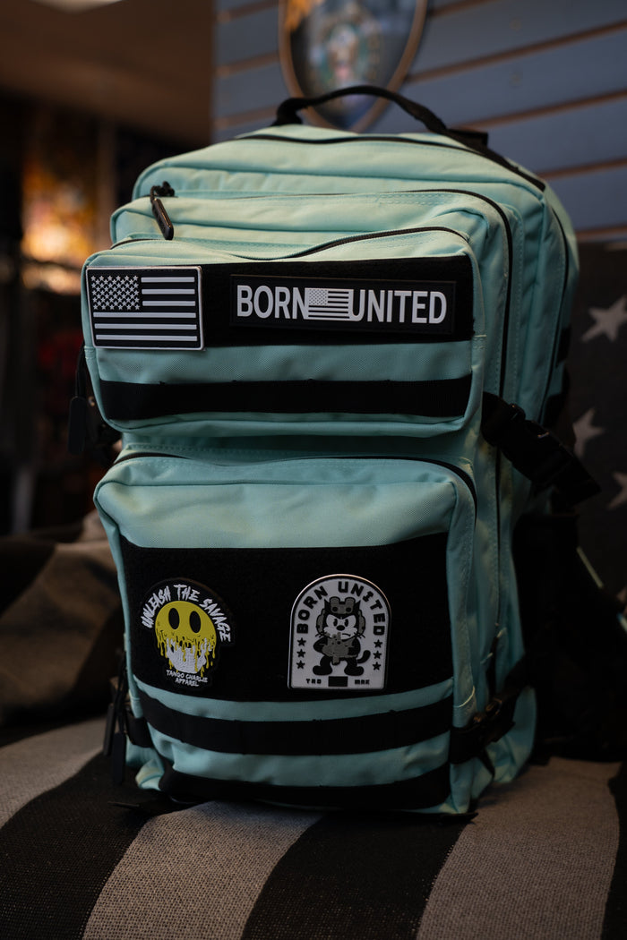 A Mint Condition Premium Tac-Pac - 45L travel backpack from Born United Private Label, shown indoors, comes in light blue and features black-and-white “BORN UNITED” and “ALONG THE WAKE” patches, along with a patch depicting a cartoon character.