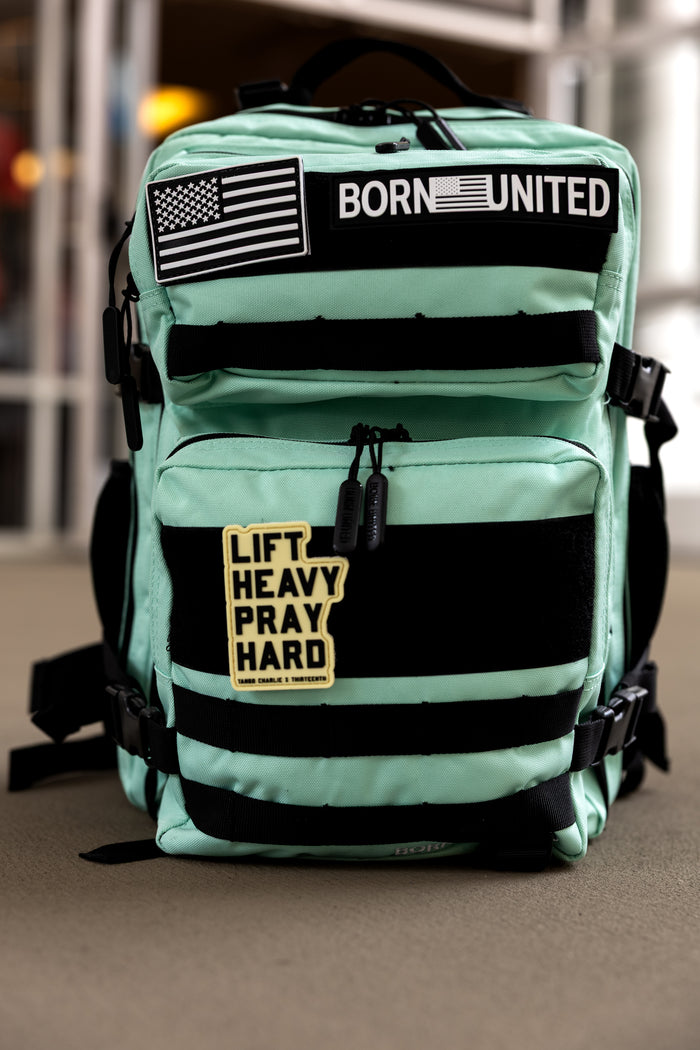 The Mint Condition Tac-Pac - 25L by Born United Private Label is a mint green backpack with black straps and a modular patch panel displaying BORN UNITED, an American flag, and LIFT HEAVY PRAY HARD patches on the front.