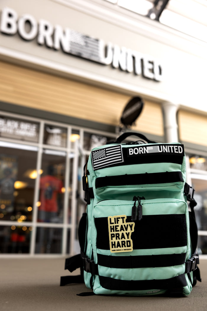 The Mint Condition Tac-Pac - 25L by Born United Private Label, a mint green tactical backpack with black straps, modular patch panel, "BORN UNITED" and flag patch, plus "LIFT HEAVY PRAY HARD" patch, sits before a store marked BORN UNITED.