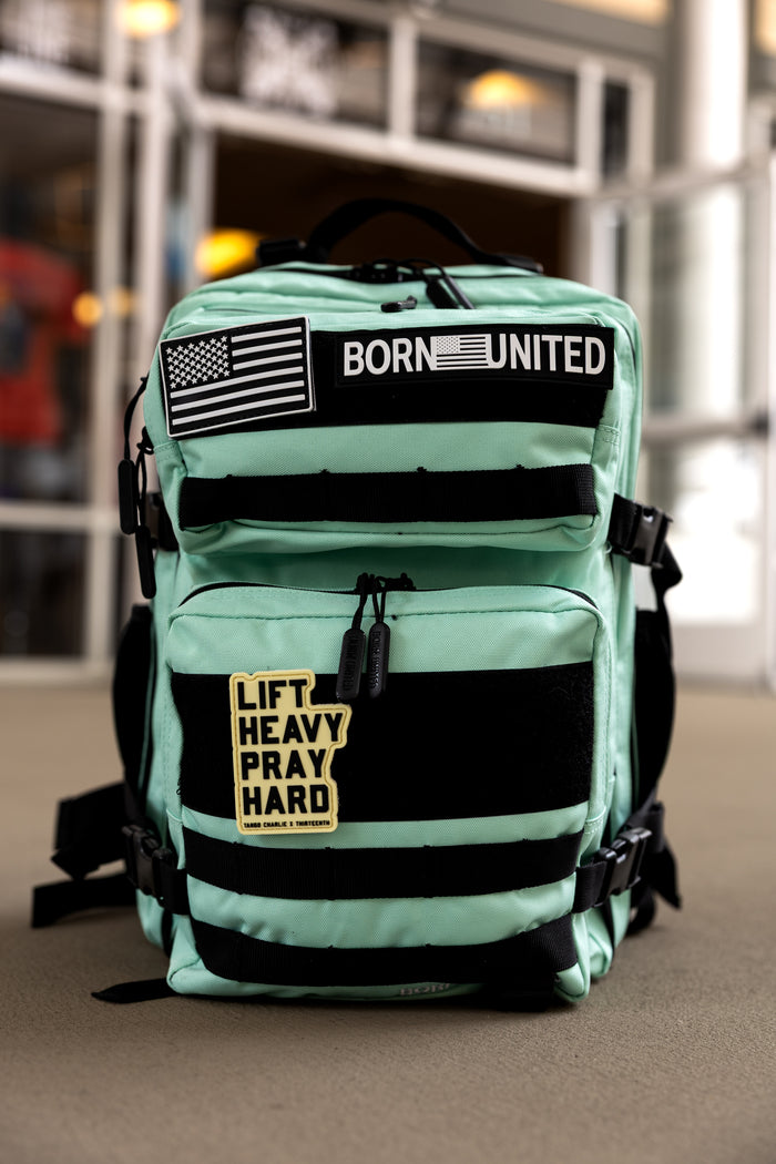 The Mint Condition Tac-Pac - 25L by Born United Private Label, featuring black straps and a modular patch panel with BORN UNITED, two American flags, and a LIFT HEAVY PRAY HARD patch, is shown indoors on the floor.