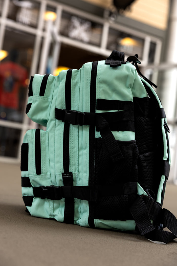 The Mint Condition Tac-Pac - 25L by Born United Private Label, featuring mint green and black colors, multiple pockets, organized storage, and a modular patch panel, stands upright on a brown surface with windows and indoor lighting in the background.