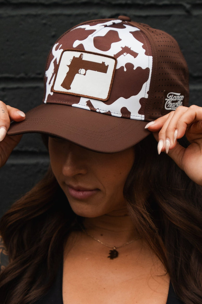 A woman in a black sports bra and the Tango Charlie Apparel Moo Moo Pew Hydro Snapback Hat, featuring a camo pattern and gun graphic, stands against a black brick wall. Her tattooed arms show as she holds the brim, face mostly hidden by the hat.