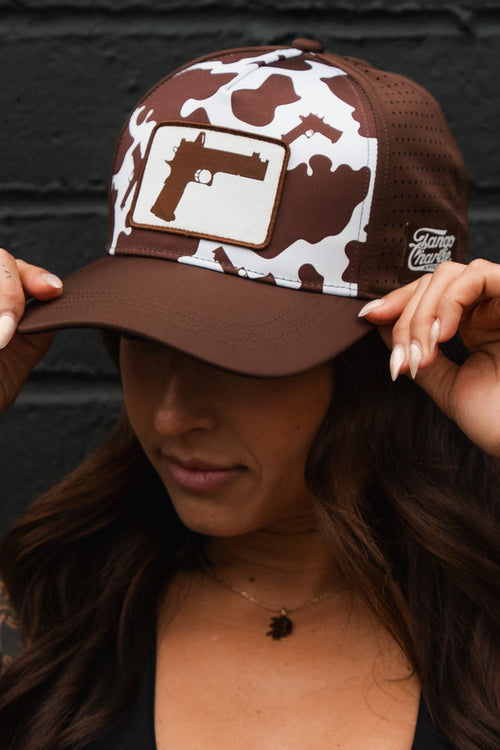 A woman in a black sports bra and the Tango Charlie Apparel Moo Moo Pew Hydro Snapback Hat, featuring a camo pattern and gun graphic, stands against a black brick wall. Her tattooed arms show as she holds the brim, face mostly hidden by the hat.