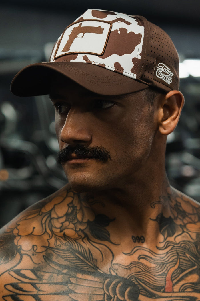 A mustached man with chest tattoos looks to the side, wearing Tango Charlie Apparel's Moo Moo Pew Hydro snapback—a brown and white camo cap featuring a stylized F and a Franco Flame patch.