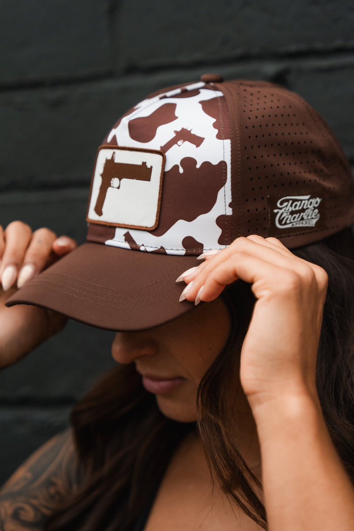 A person wearing the Moo Moo Pew Hydro Snapback Hat by Tango Charlie Apparel, featuring a brown and white camo pattern, hydro resistance, a gun silhouette patch on the front, and "Tango Charlie" text on the side.