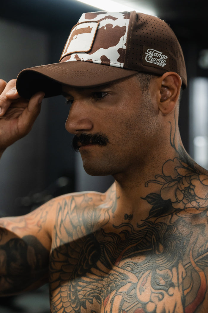 A man with a mustache and tattoos adjusts a Moo Moo Pew Hydro - Snapback Hat by Tango Charlie Apparel while standing indoors.