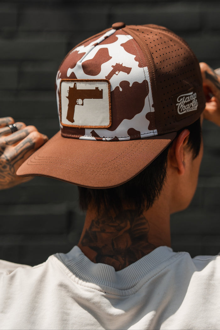 A person with hand and neck tattoos adjusts the Tango Charlie Apparel Moo Moo Pew Hydro snapback hat, featuring a brown and white camouflage pattern, breathable mesh, and gun graphics.