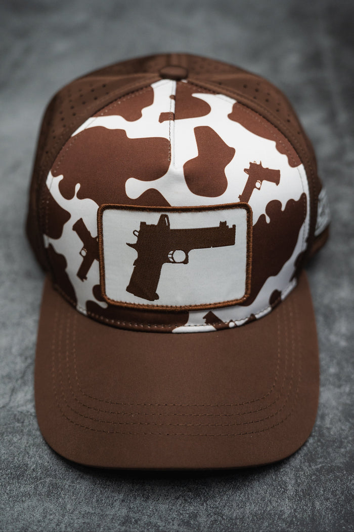 The Moo Moo Pew Hydro Snapback Hat by Tango Charlie Apparel is a handmade brown and white cap with a camouflage gun silhouette pattern and large front gun image, shown on a gray surface.