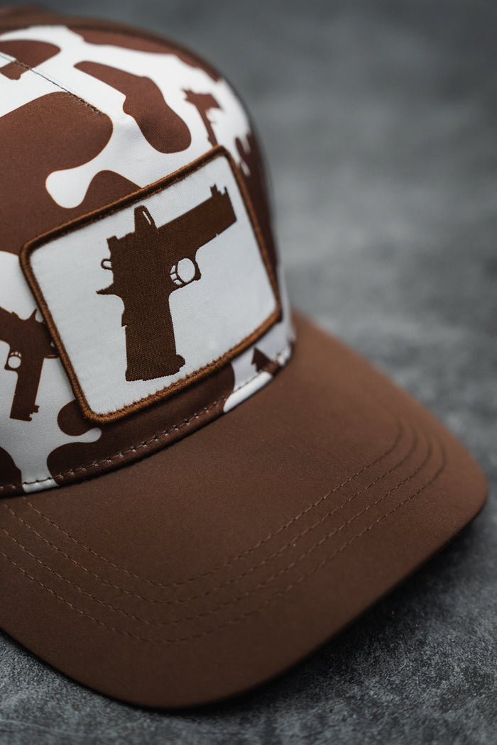 The Moo Moo Pew Hydro Snapback Hat by Tango Charlie Apparel features brown and white camouflage, a breathable design, and a handgun silhouette patch on the front, shown on a gray textured surface.