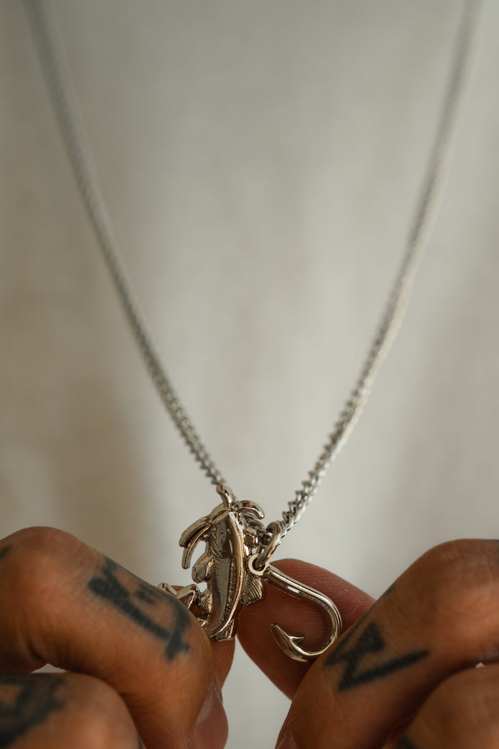 A person with tattooed fingers holds the Mud Cat Noodler - Stainless Steel Necklace by Mucker Bucket, which features a stainless steel fish and hook pendant suspended from a matching chain, displayed against a plain white shirt background.
