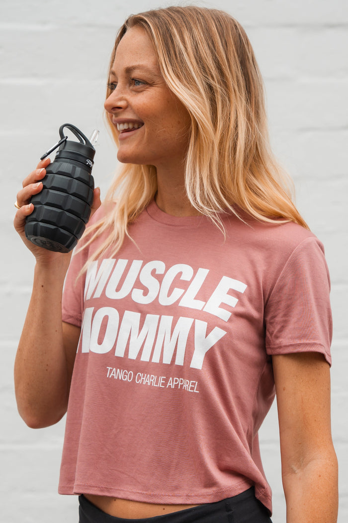A fit, smiling woman with blonde hair wears the Muscle Mommy - Crop Tee by Tango Charlie Apparel and holds a black grenade-shaped water bottle—a true muscle mommy who can deadlift 225 lbs.