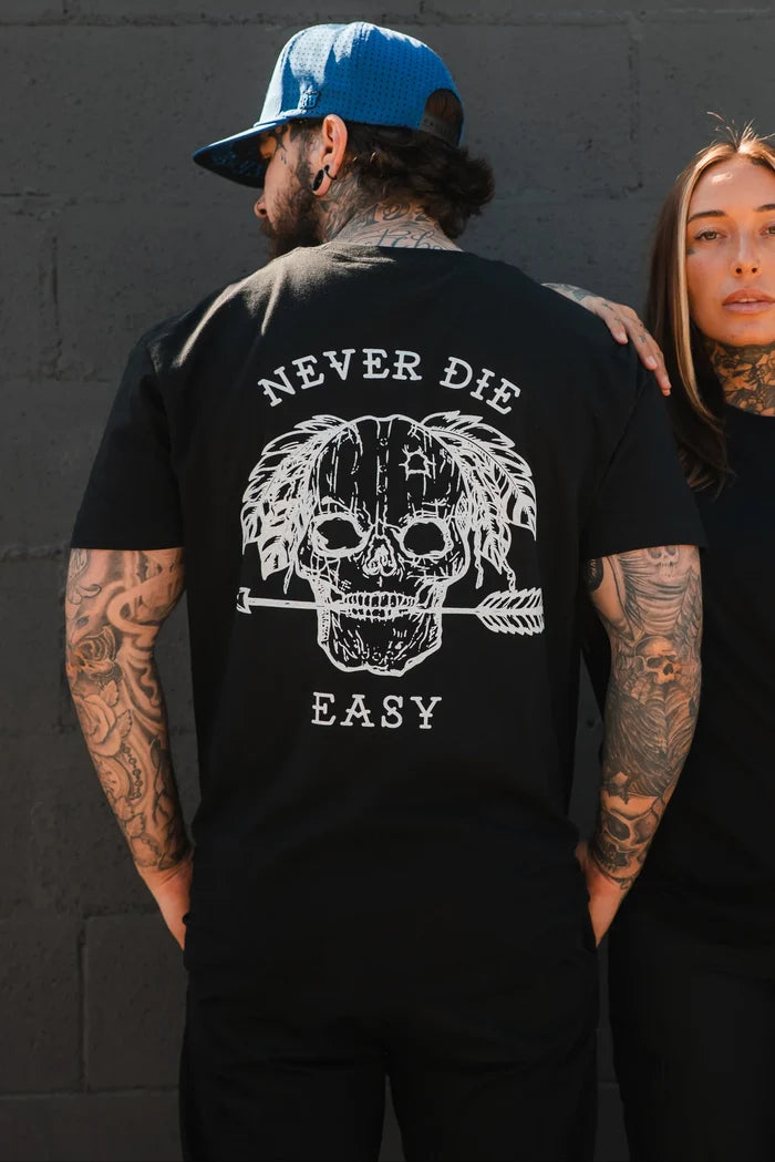 A tattooed man wears the Relentless Betrayal Never Die Easy Black Tee, made from fine jersey cotton and featuring a skull, feathers, an arrow, and "NEVER DIE EASY" on the back, standing next to a tattooed woman by a dark brick wall.