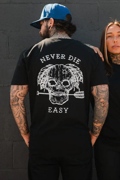 A tattooed man wears the Relentless Betrayal Never Die Easy Black Tee, made from fine jersey cotton and featuring a skull, feathers, an arrow, and "NEVER DIE EASY" on the back, standing next to a tattooed woman by a dark brick wall.