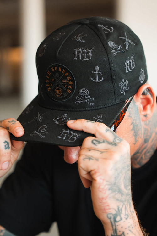 A tattooed individual adjusts the brim of a Relentless Betrayal Never Surrender Limited Edition Snapback, featuring white symbols and a circular patch. Their face remains mostly hidden.