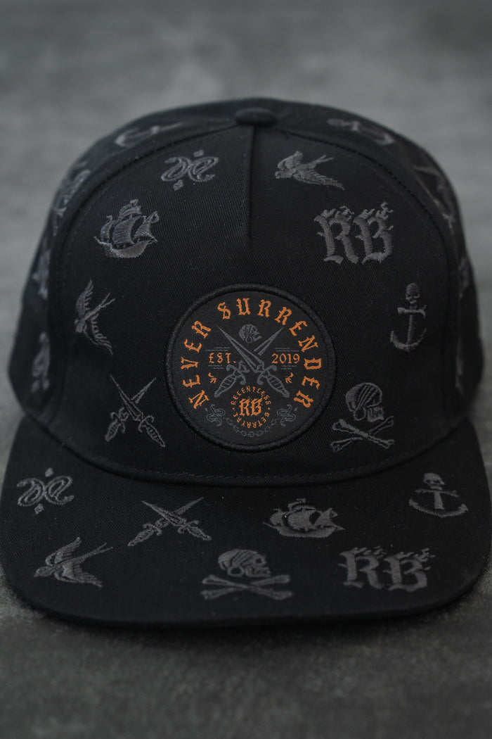 The Never Surrender Limited Edition Snapback by Relentless Betrayal is a black cap with grey nautical and tattoo-style designs, plus an orange and black patch on the front reading "Never Surrender Est. 2019.