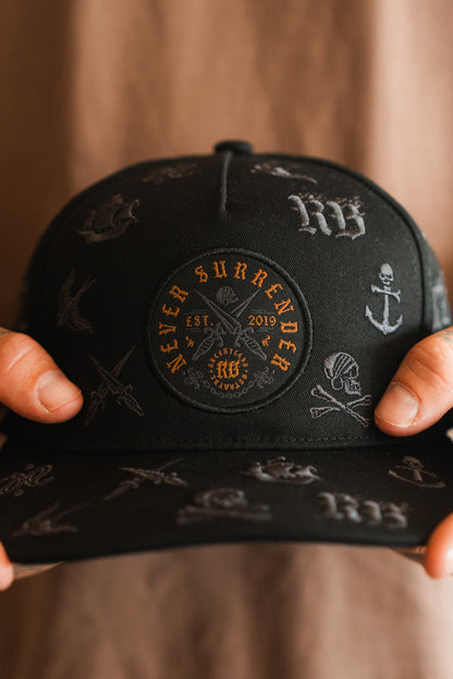 A person holds the Relentless Betrayal Never Surrender Limited Edition Snapback, featuring embroidered skulls, swords, anchors, and a circular patch with "Never Surrender" and "Est. 2019"—a bold choice for collectors.