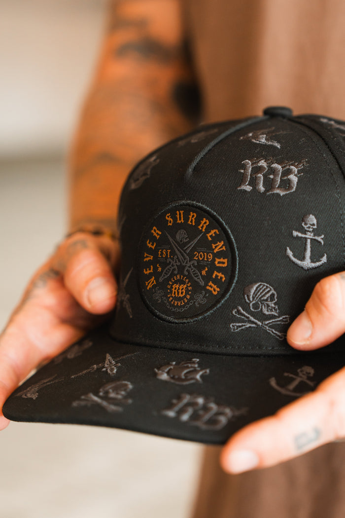 A tattooed hand holds the Relentless Betrayal Never Surrender Limited Edition Snapback, a black cap featuring embroidered nautical symbols and a circular patch stating NEVER SURRENDER EST. 2019.