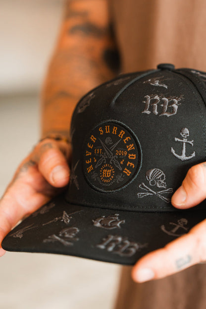 A tattooed hand holds the Relentless Betrayal Never Surrender Limited Edition Snapback, a black cap featuring embroidered nautical symbols and a circular patch stating NEVER SURRENDER EST. 2019.