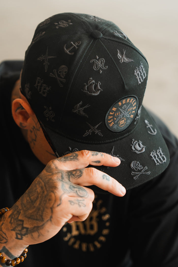 A person with tattooed hands wears the Never Surrender Limited Edition Snapback by Relentless Betrayal—a black cap with white embroidered symbols and a circular EVER SINCE patch—tilting it to partially cover their face.